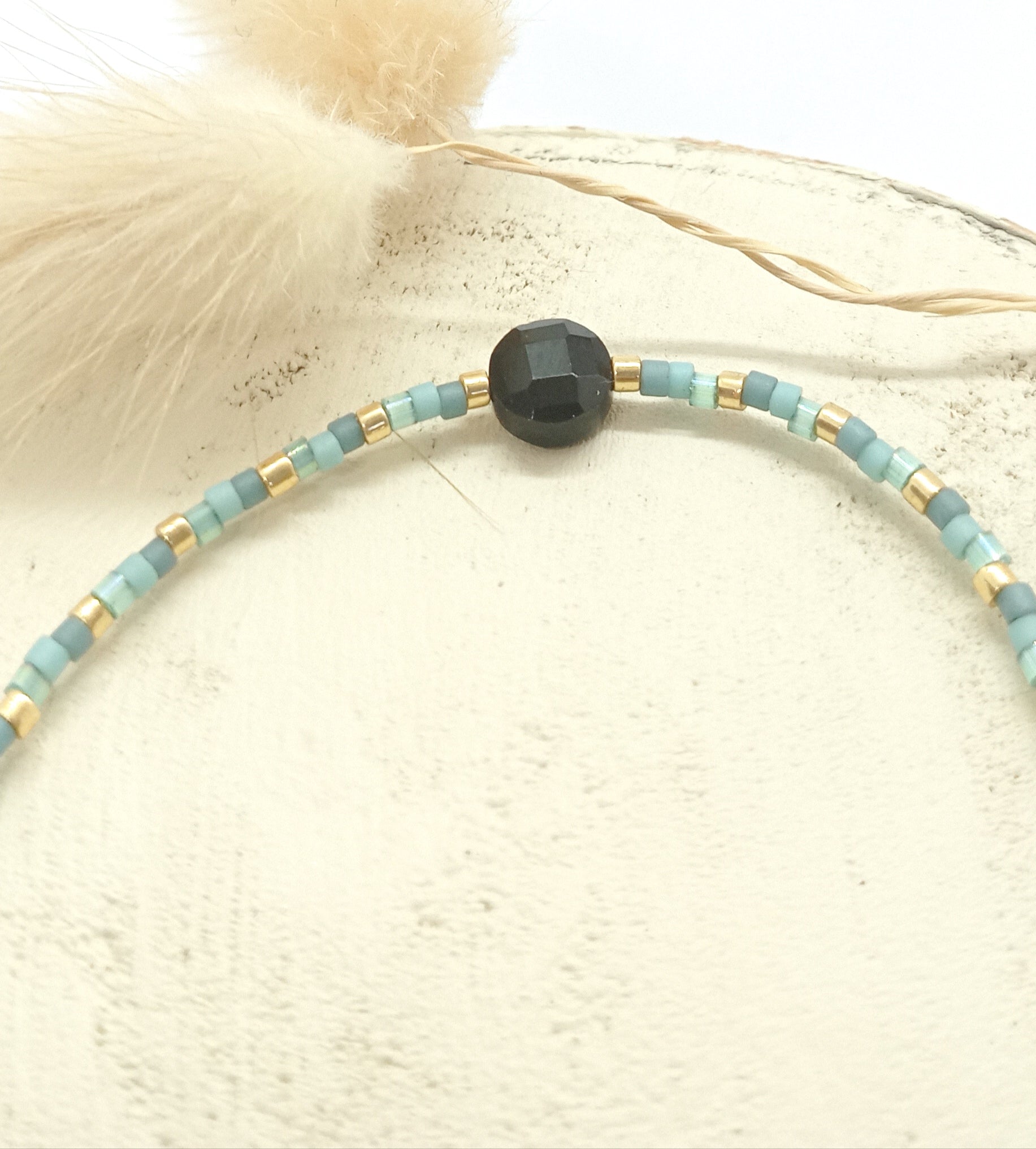 Bracelet en perles bleues et pierre noire sur un rond de bois écru et fleurs séchées en arrière plan. 