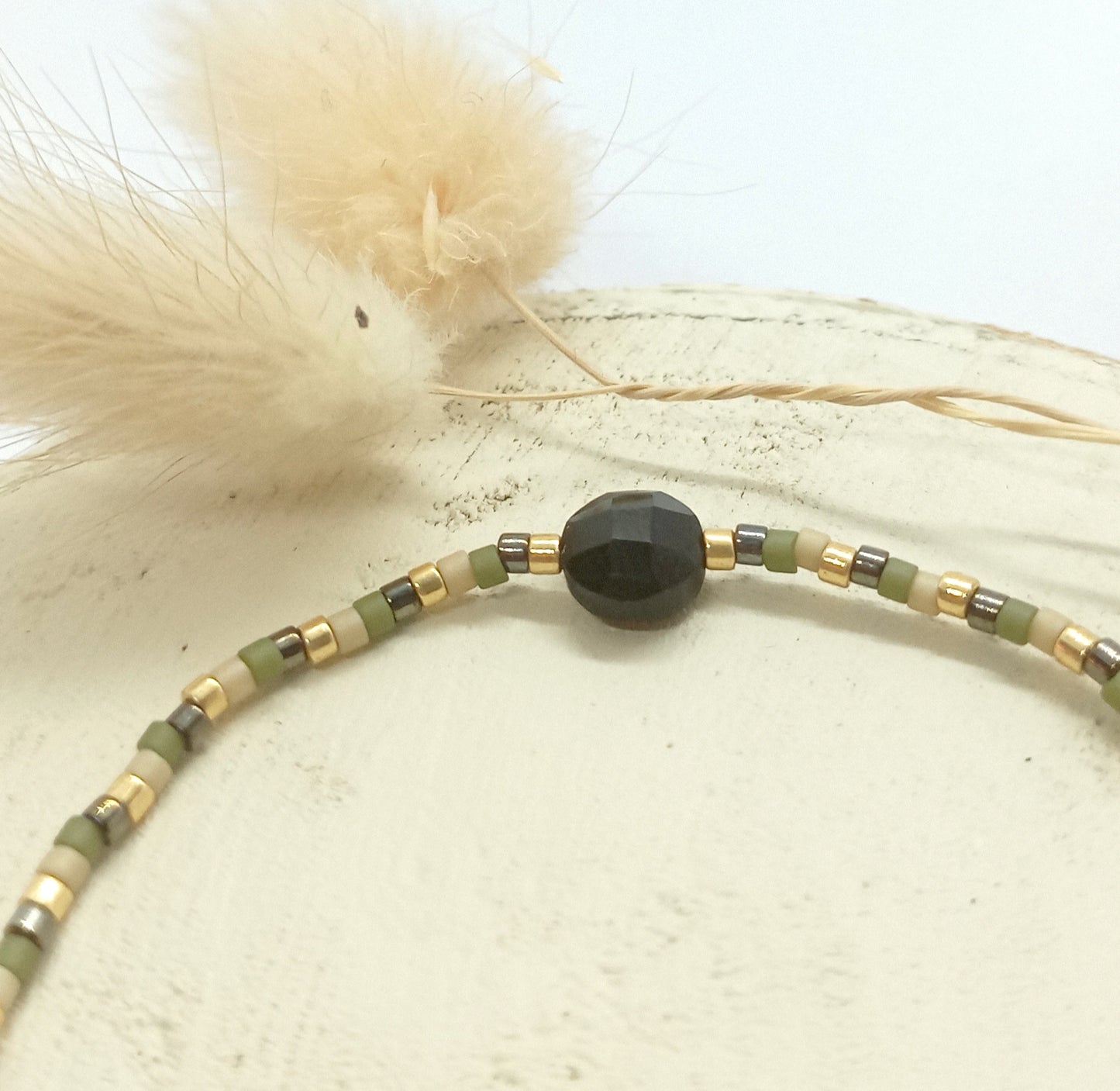 Bracelet en perles kaki et pierres Onyx noire posé sur un rond de bois écru et fleurs séchées en arrière plan.