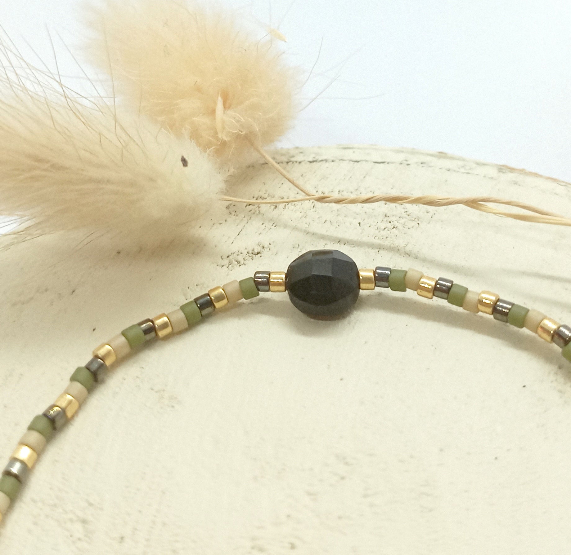 Bracelet en perles kaki et pierres Onyx noire posé sur un rond de bois écru et fleurs séchées en arrière plan. 