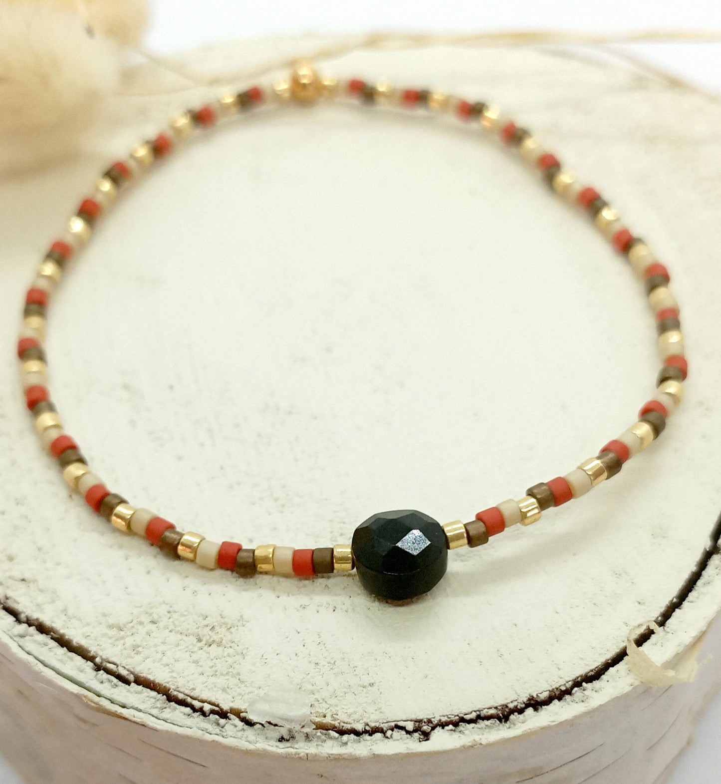 Bracelet en perles miyuki terre, beige et doré avec une pierre centrale en onyx noir. Le bracelet est posé sur un rond de bois écru, en arrière plan des fleurs séchées.