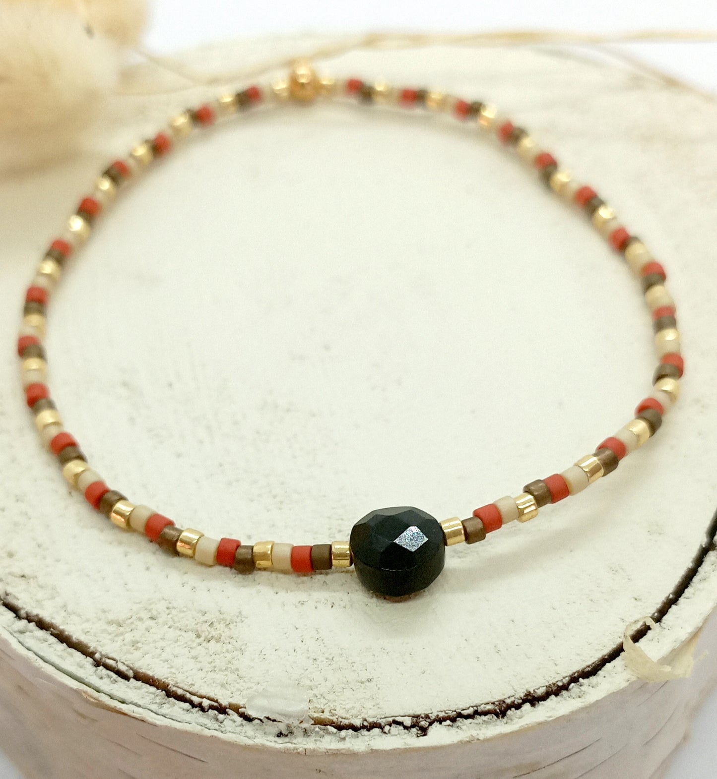 Bracelet en perles miyuki terre, beige et doré avec une pierre centrale en onyx noir. Le bracelet est posé sur un rond de bois écru, en arrière plan des fleurs séchées. 