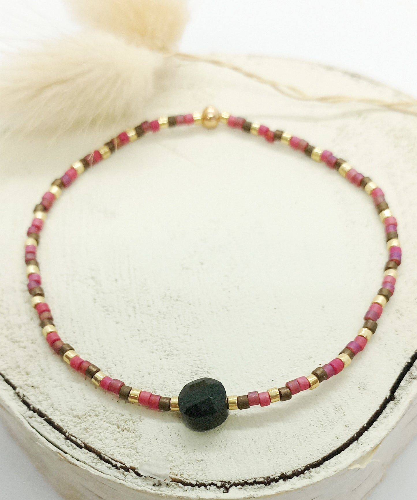 Bracelet en perles miyuki roses, marrons et dorées avec une pierre centrale en onyx. Le bracelet est posé sur un rond de vois écru. Des fleurs séchées en arrière plan.