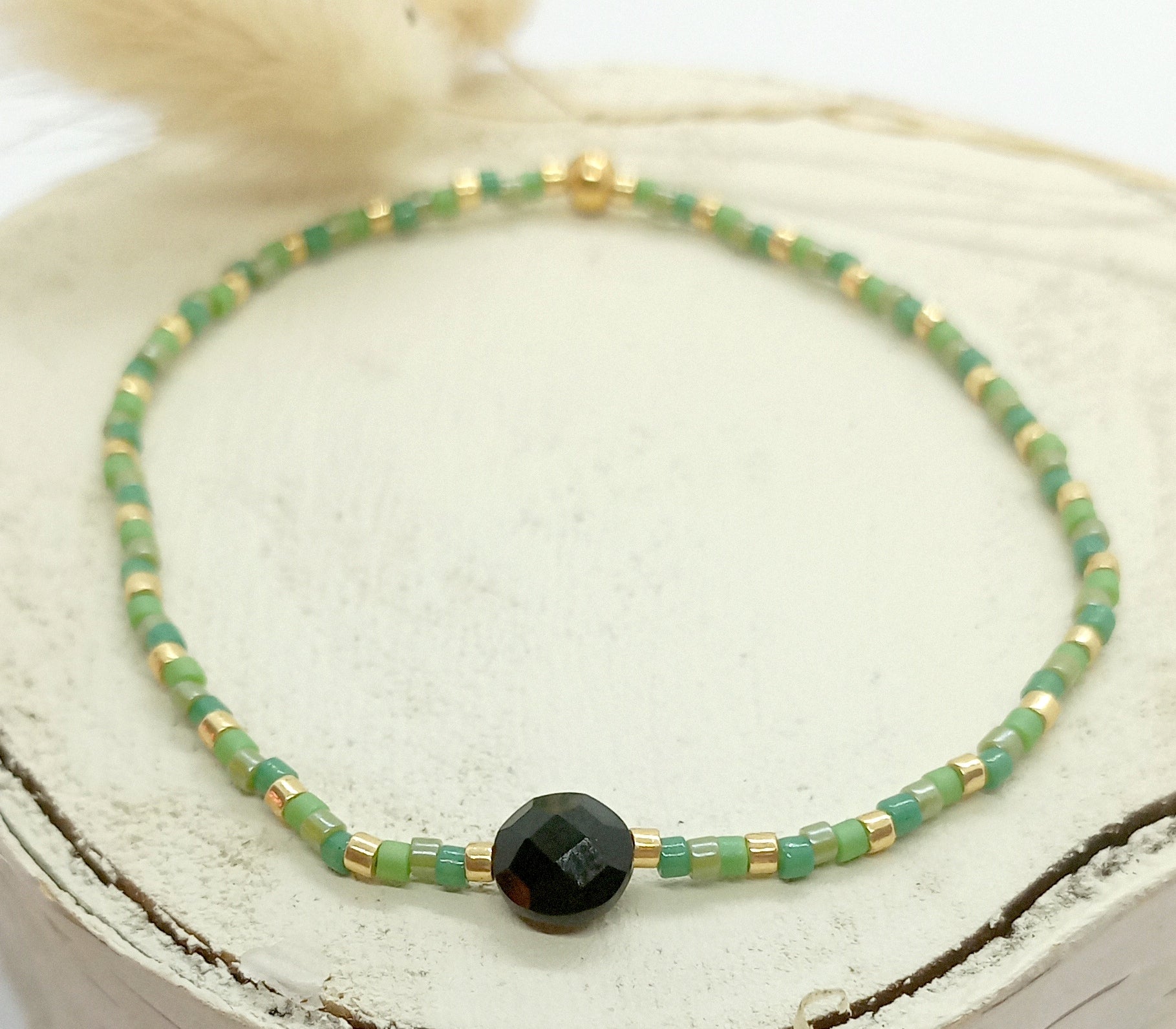 Bracelet en perles miyuki vertes et dorées avec une pierre centrale en onyx. Le bracelet est posé sur un rond de vois écru. Des fleurs séchées en arrière plan.