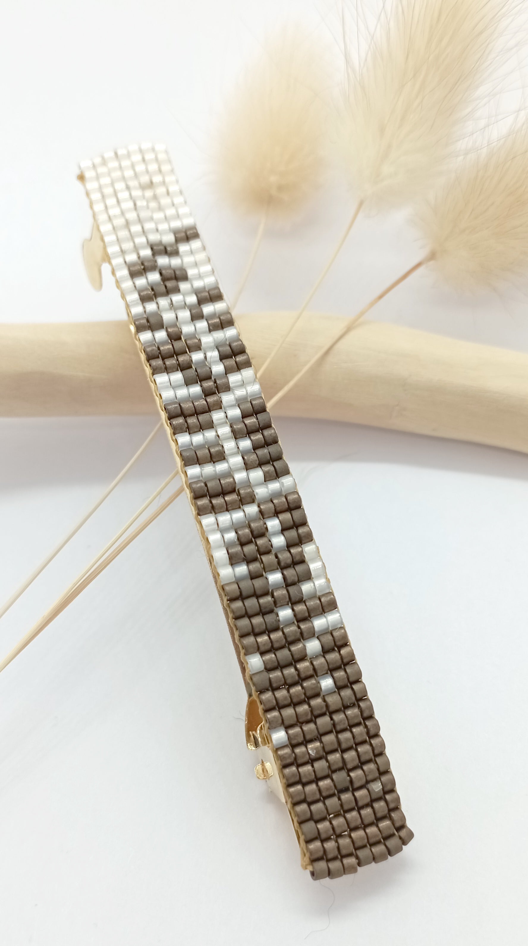 Barrette en métal et en perles tissées marron et blanche sur un fond blanc, une branche de bois et des fleurs séchées. 