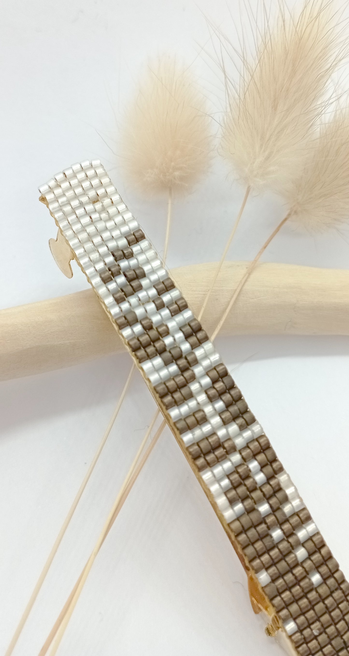 Barrettes en métal et en perles tissées marron et blanches posée sur trois fleurs séchées beiges.