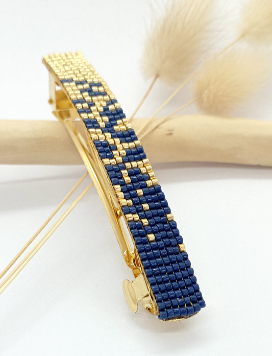 Barrette pour cheveux en métal doré et perles tissées bleue marine et dorée. sur un fond blanc et une branche de bois clair.