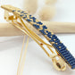 Barrette pour cheveux en métal dorée et en perles tissées bleues marine et dorées sur un fond blanc et une branche de bois clair.  