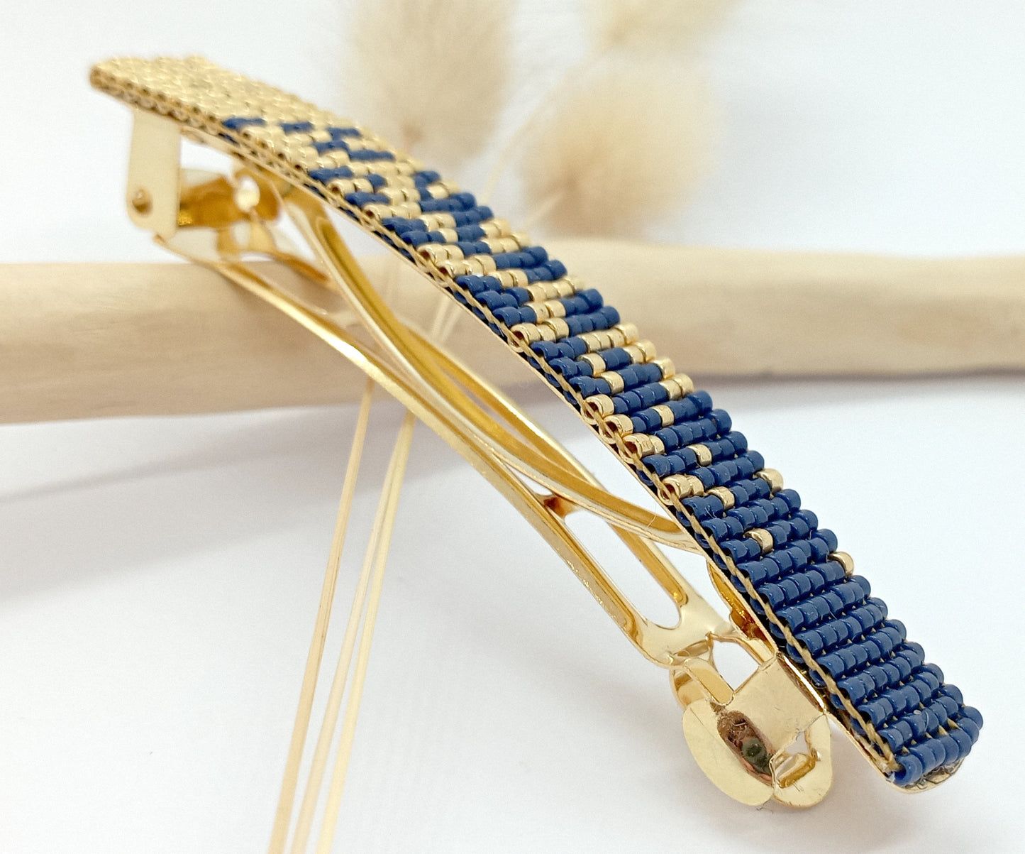 Barrette pour cheveux en métal dorée et en perles tissées bleues marine et dorées sur un fond blanc et une branche de bois clair.