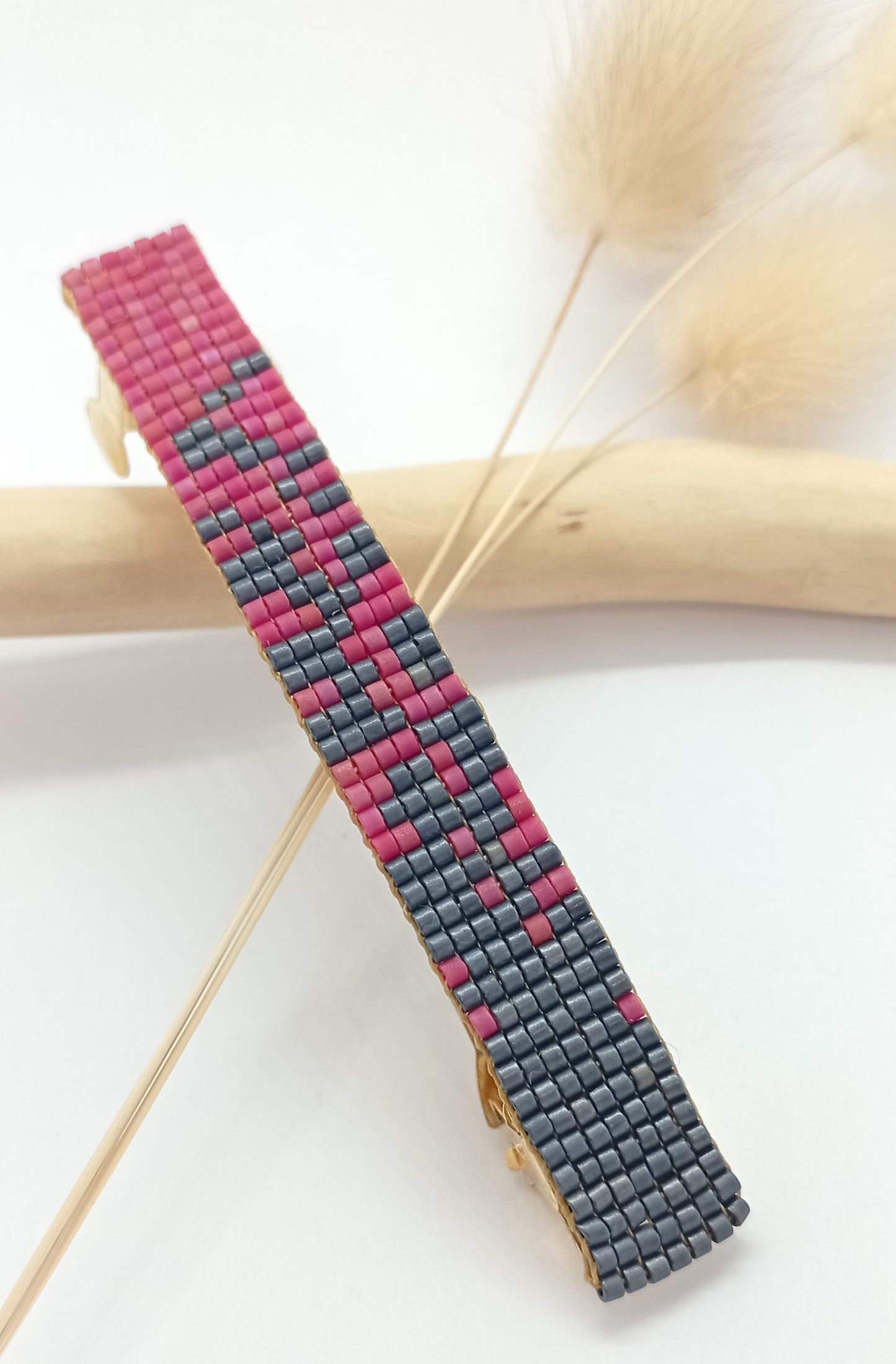 Barrette pour cheveux en perles tissées grises et roses sur un fond blanc et une branche de bois clair.
