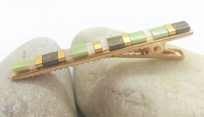 Pince crocodile en métal doré et en perles plates de couleurs vertes, dorées et marrons, posée sur deux cailloux et un fond blanc. 