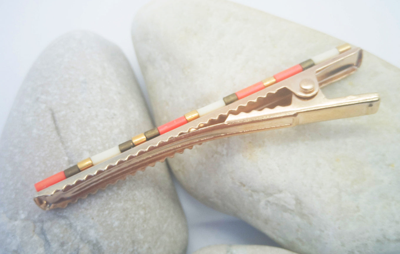 Barrette pour cheveux type pince crocodile avec dent en métal doré posé sur des cailloux et un fond blanc.