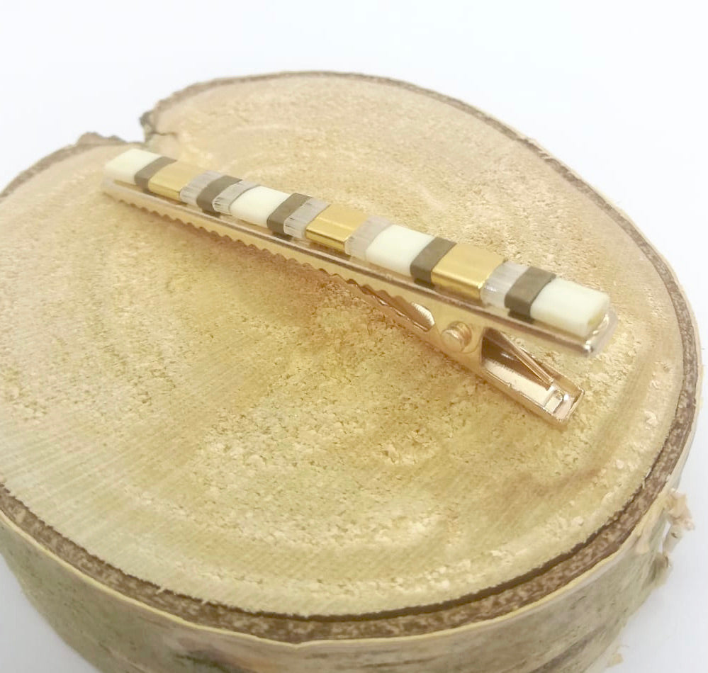 Barrette pour cheveux type pince crocodile en métal doré et perles plates écru, posée sur un rond de bois et un fond blanc.