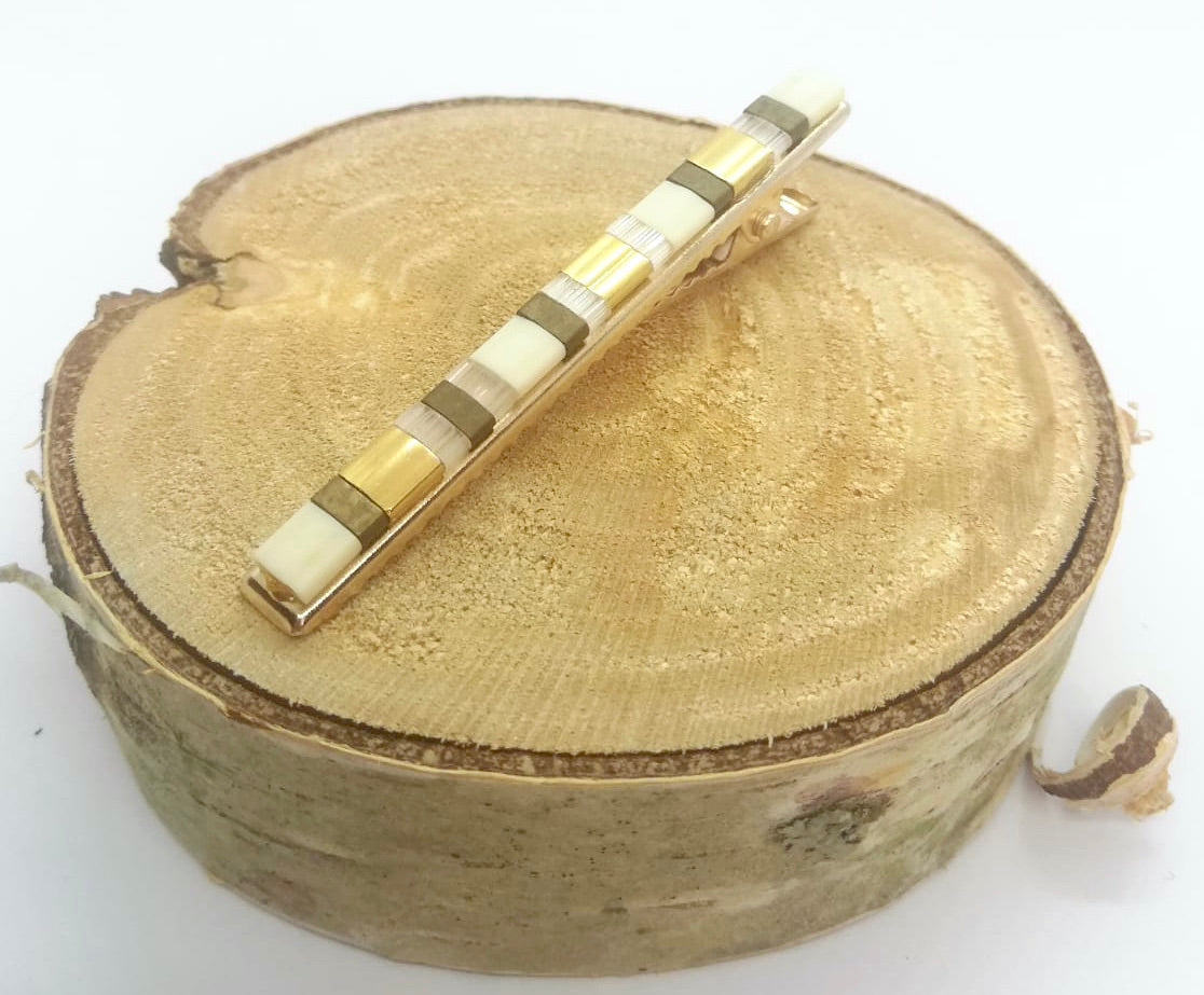 Barrette pour cheveux type pince crocodile en métal doré et perles plate, posée sur un rond de bois et un fond blanc. 