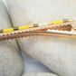Pince crocodile en métal doré avec dents, posée sur des cailloux et un fond blanc. 