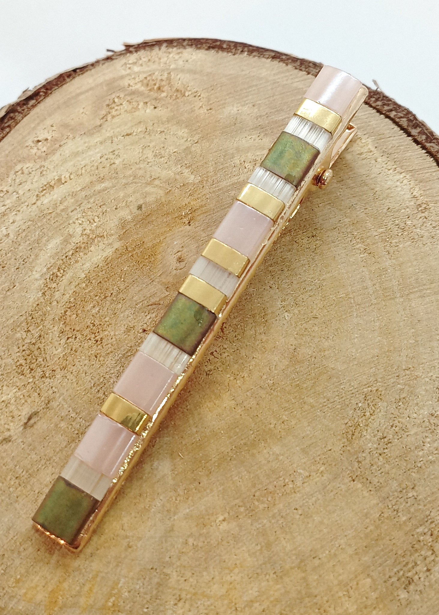 Barrette pour cheveux en métal doré et perles plates colorées, posée sur un rond de bois et un fond blanc. 