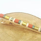 Barrette pour cheveux type pince crocodile en métal doré et perles plates colorées, posée sur un rond de bois et un fond blanc.