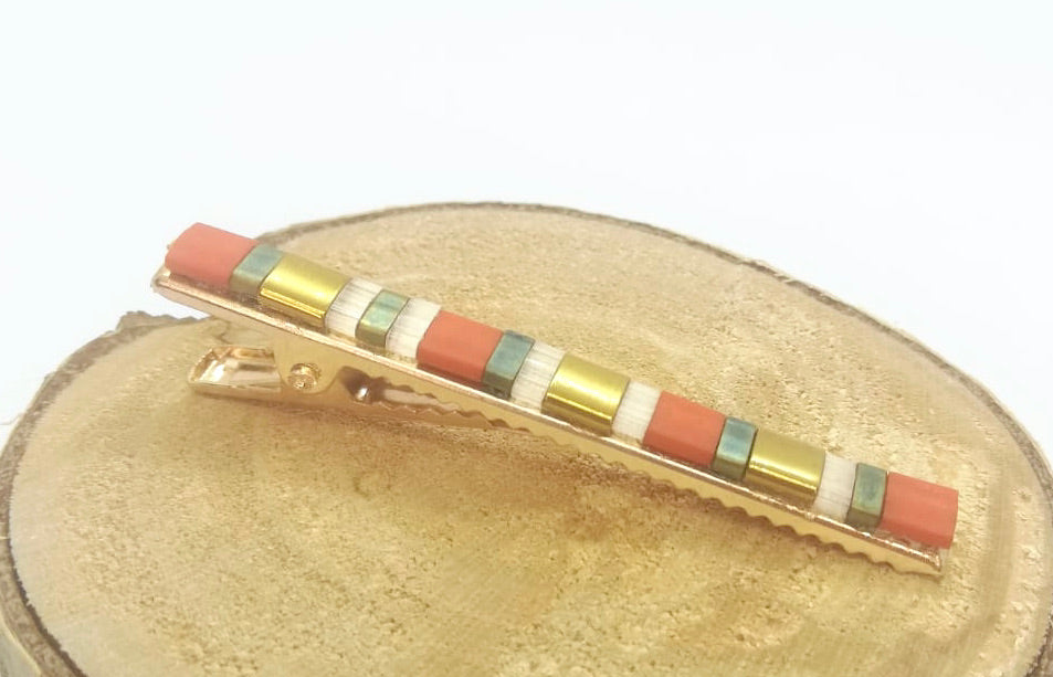 Barrette pour cheveux type pince crocodile en métal doré et perles plates colorées, posée sur un rond de bois et un fond blanc.
