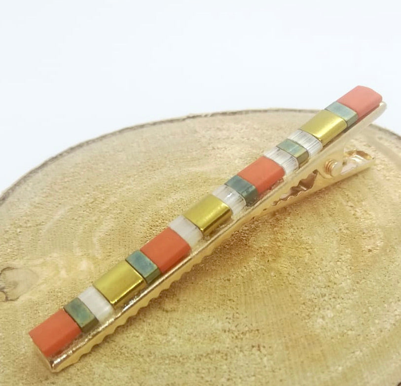 Barrette pour cheveux type pince crocodile en métal doré et perles plates de couleurs, posée sur un rond de bois et un fond blanc