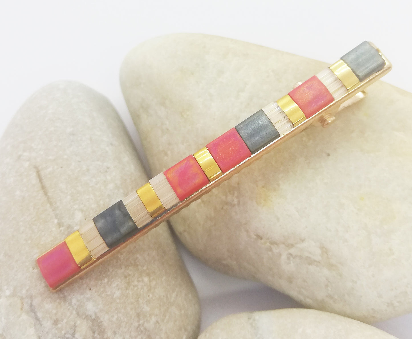 Barrette pour cheveux en métal doré et perles plates colorées, posée sur des cailloux et un fond blanc.