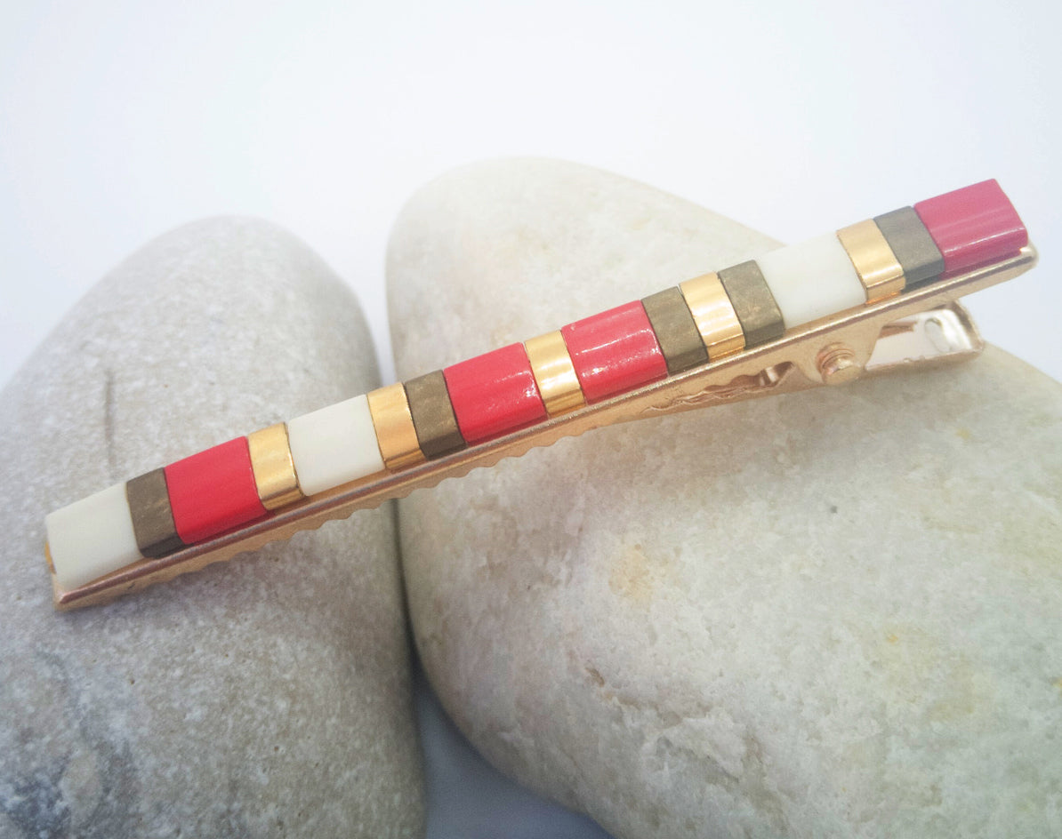 Barrette pour cheveux en métal doré et perles plates colorées, posée sur des cailloux et un fond blanc.