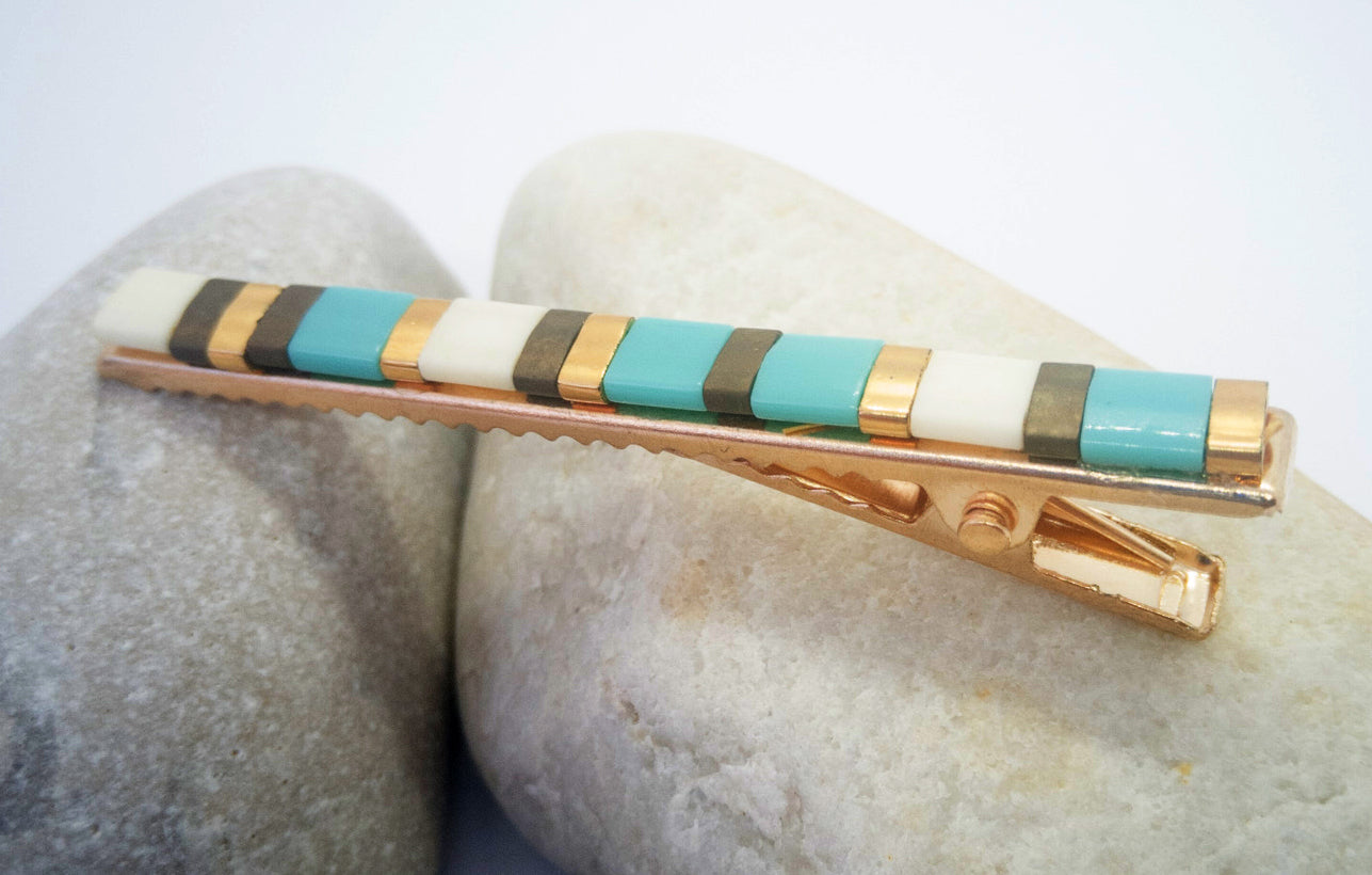 Barrette pour cheveux type pince crocodile en métal doré et perles plates de couleur, posé sur des cailloux et un fond blanc.