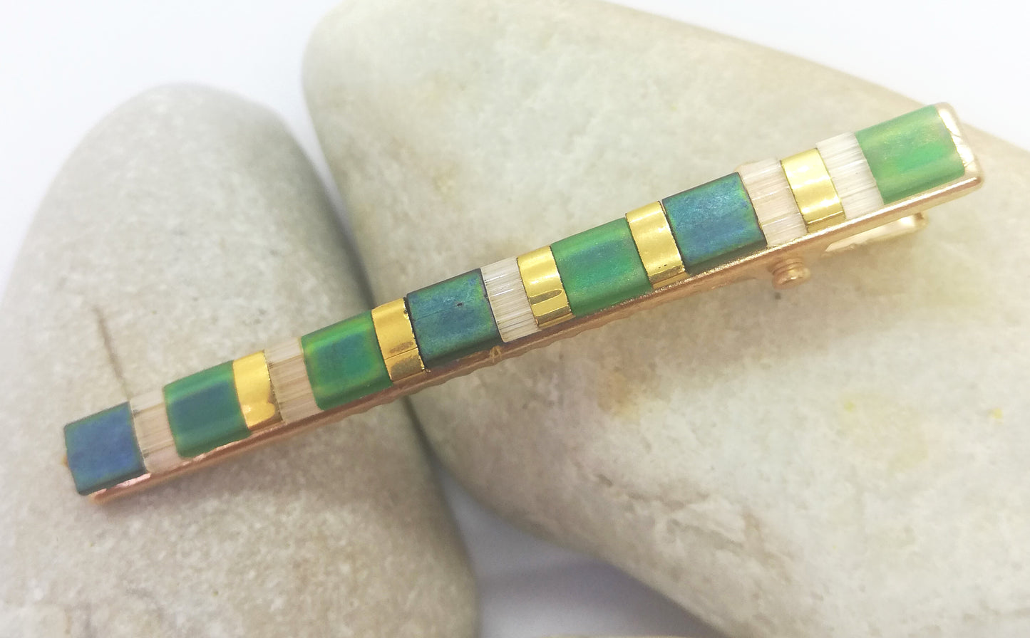 Barrette pour cheveux type pince crocodile en métal doré et perles plates de couleurs, posée sur des cailloux et un fond blanc