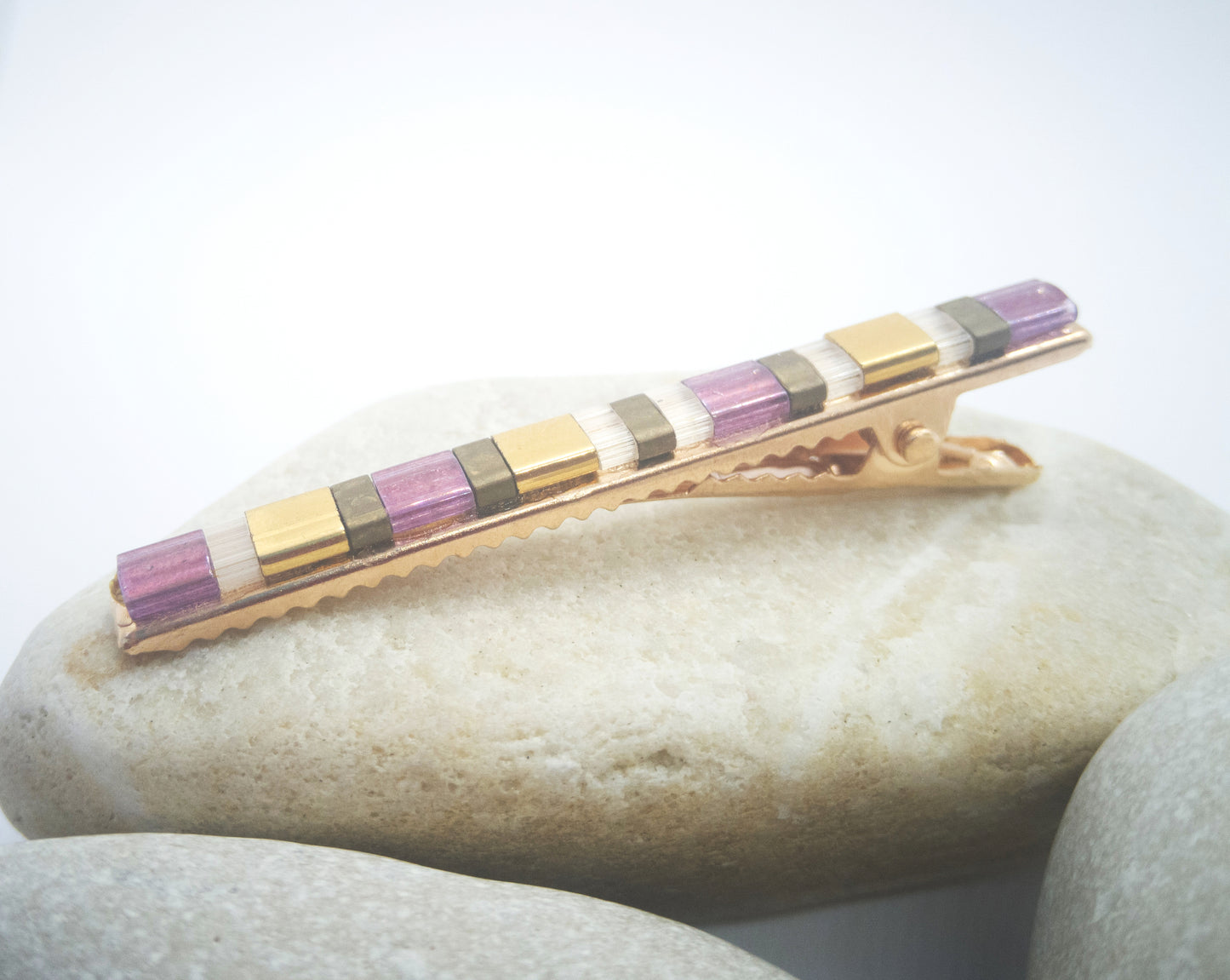 Barrette pour cheveux type pince crocodile en métal doré et perles plates de couleurs, posée sur des cailloux et un fond blanc.