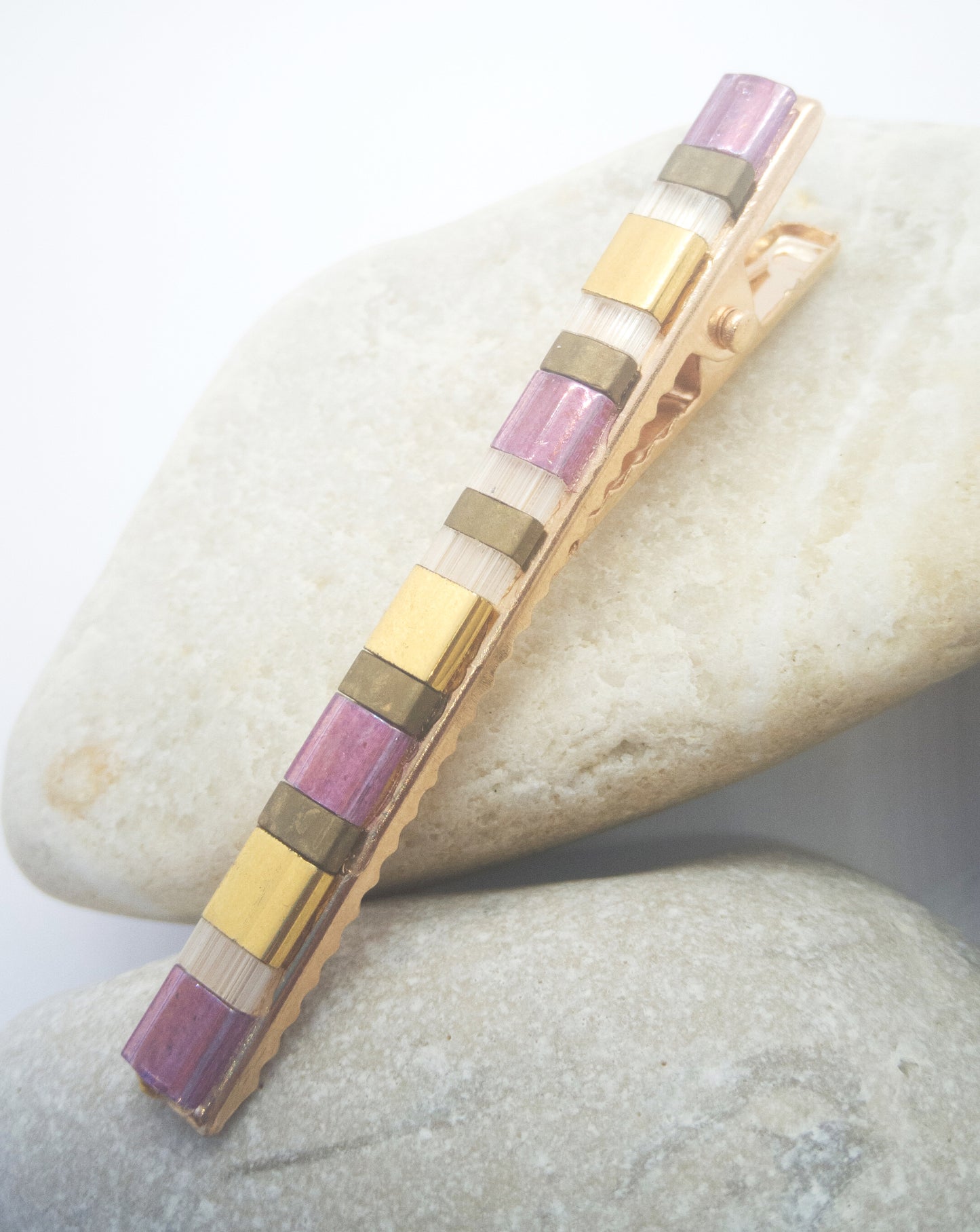 Barrette pour cheveux type pince crocodile en métal doré et perles plates de couleurs, posée sur des cailloux et un fond blanc.