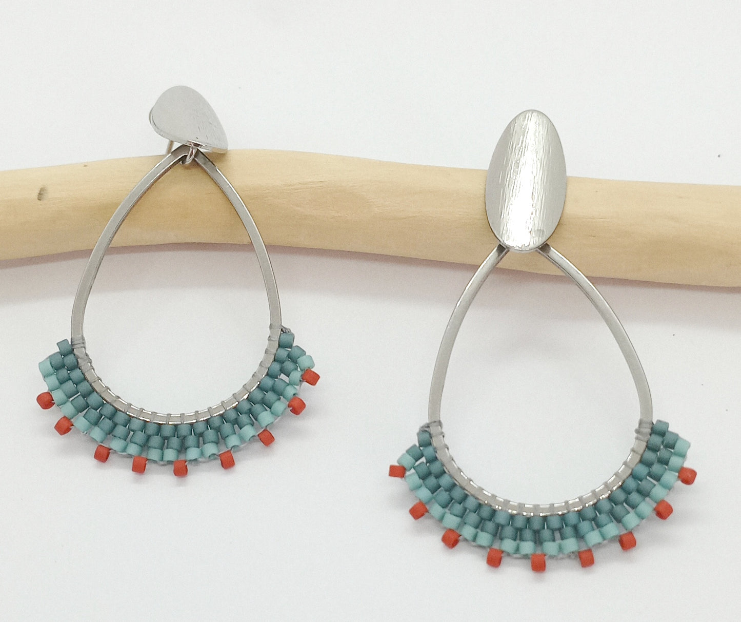 Boucles d'oreilles en forme de goutte en acier argenté et perles tissées bleues, sur un fond blanc et une branche de bois. 