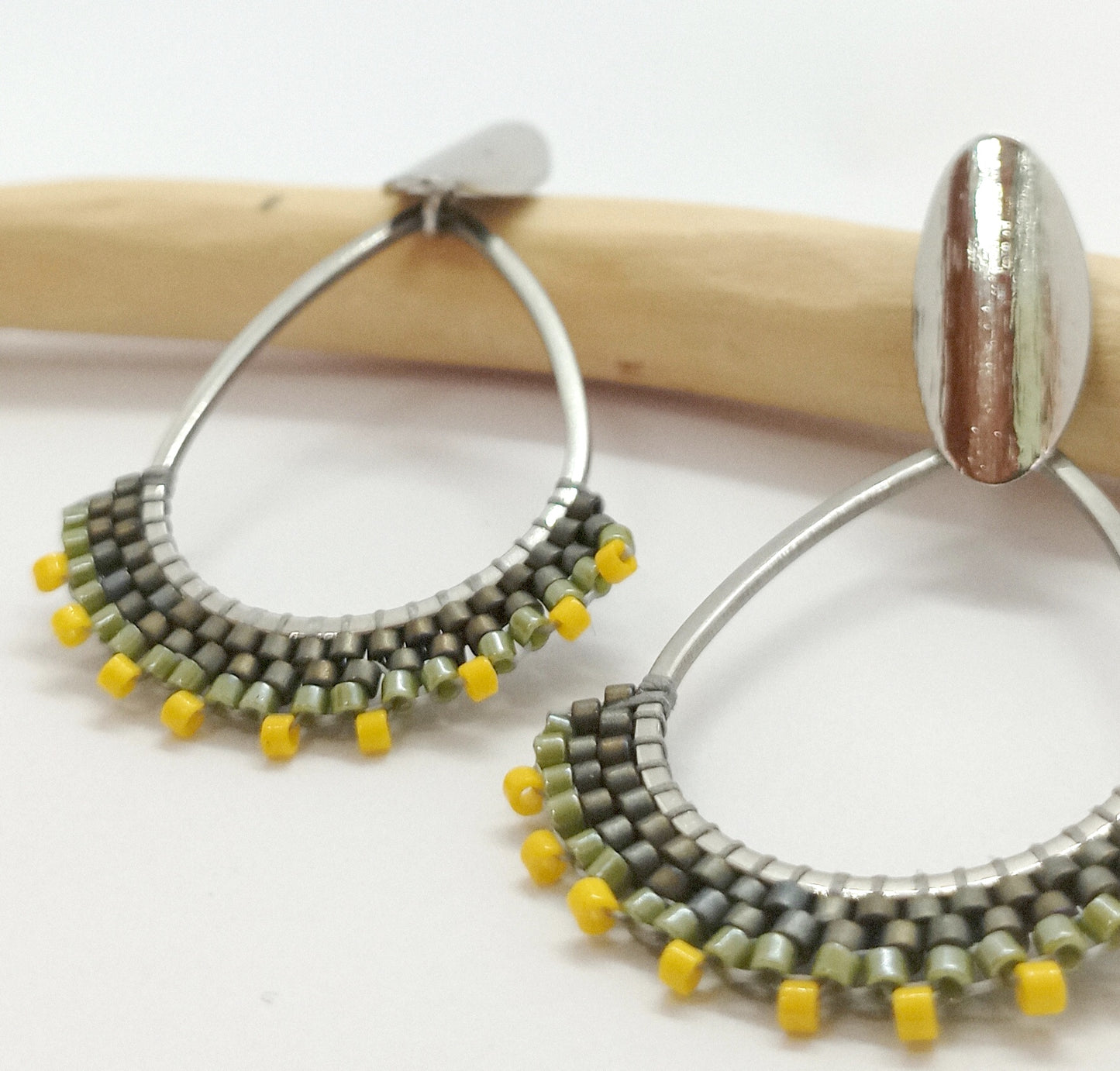 Boucles d'oreilles en forme de goutte en acier argenté et en perles tissées sur un fond blanc et une branche de bois.