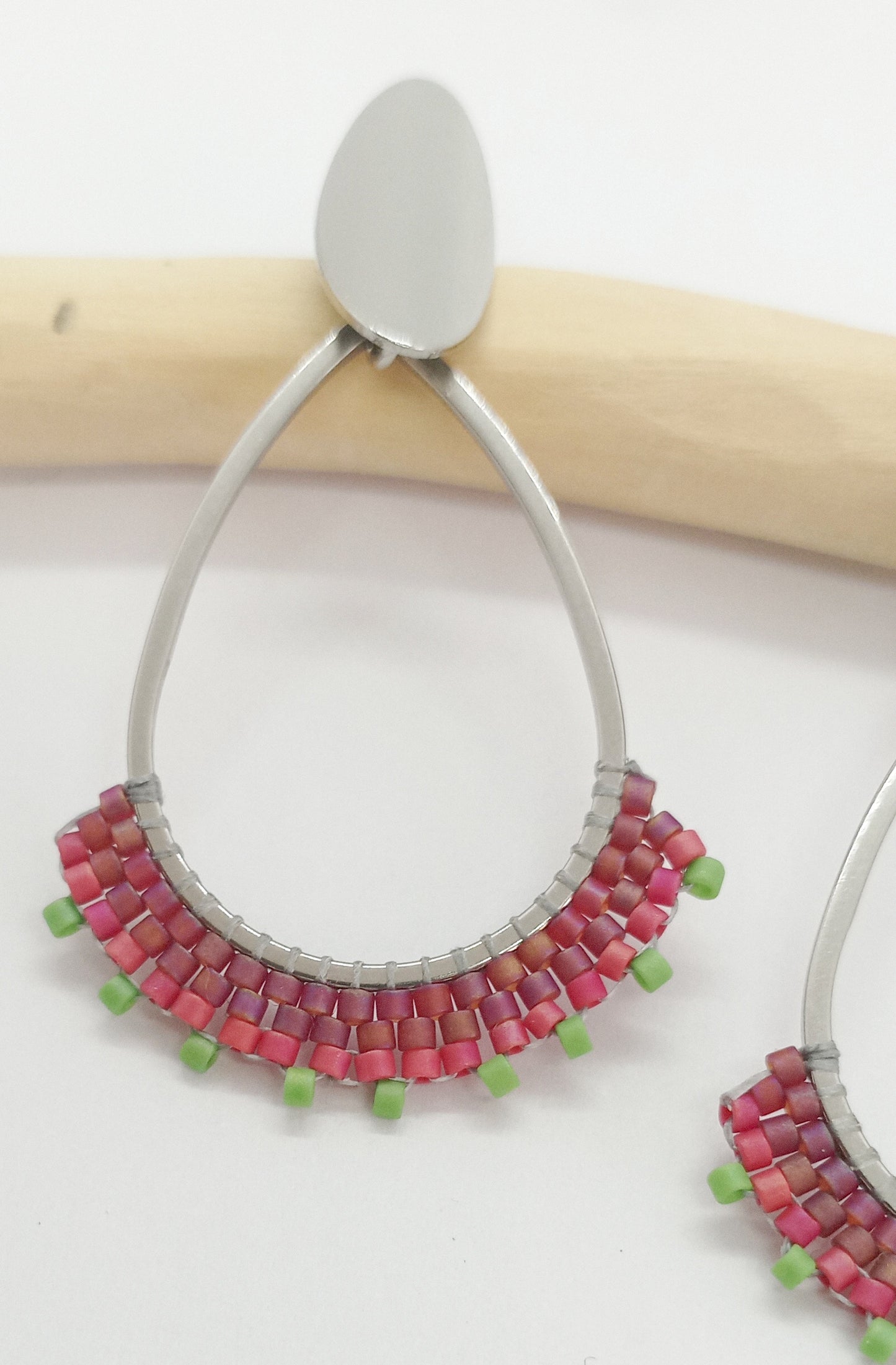 Boucles d'oreilles en acier argenté et en perles tissées rouges, posée sur une branche et un fond blanc.