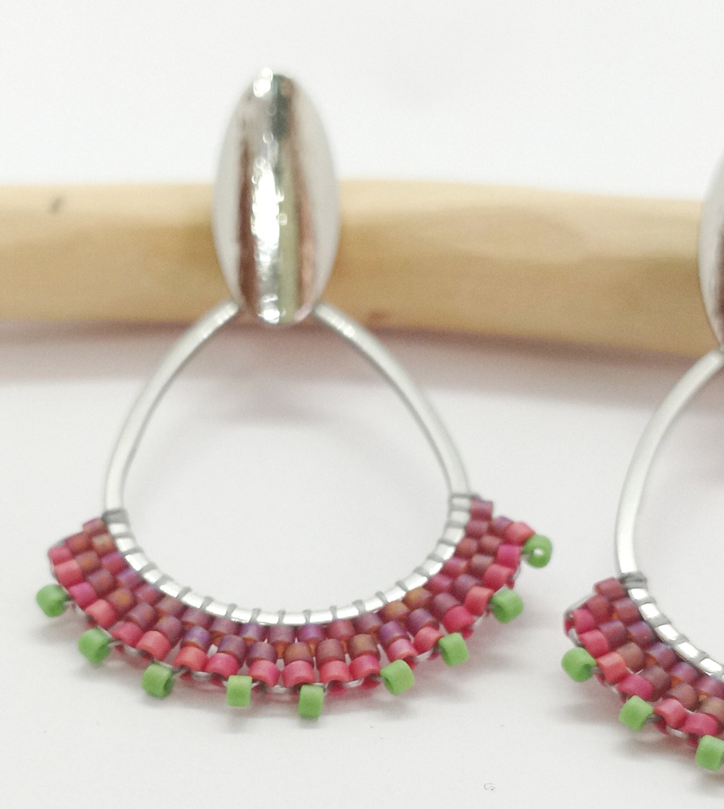 Boucles d'oreilles en acier argenté et perles miyuki tissées et posée sur une branche de bois et un fond blanc.