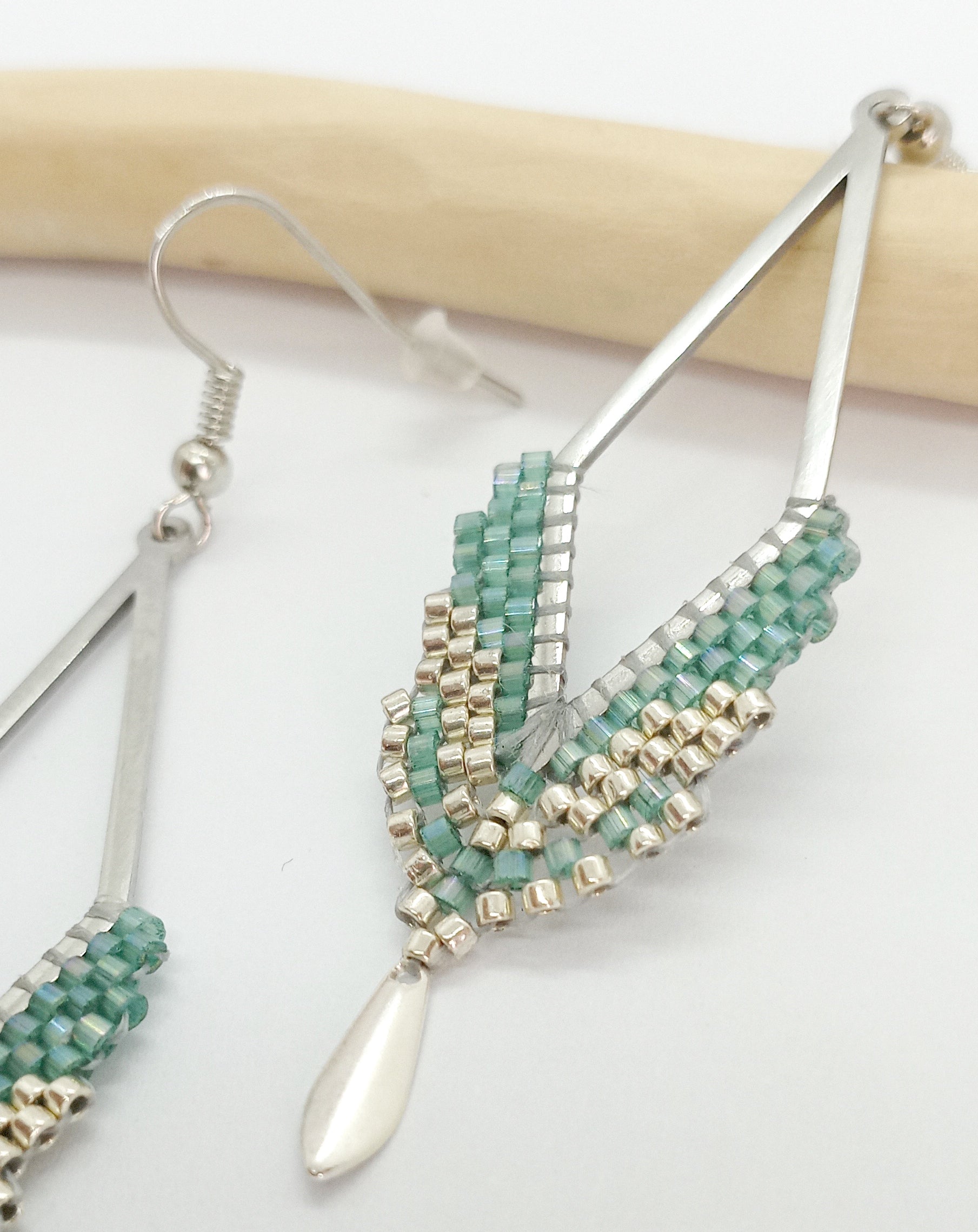 Boucles d'oreilles en acier argenté en forme de losange et perles miyuki tissées vert d'eau avec un sequin argenté. Posées sur fond blanc et bois clair