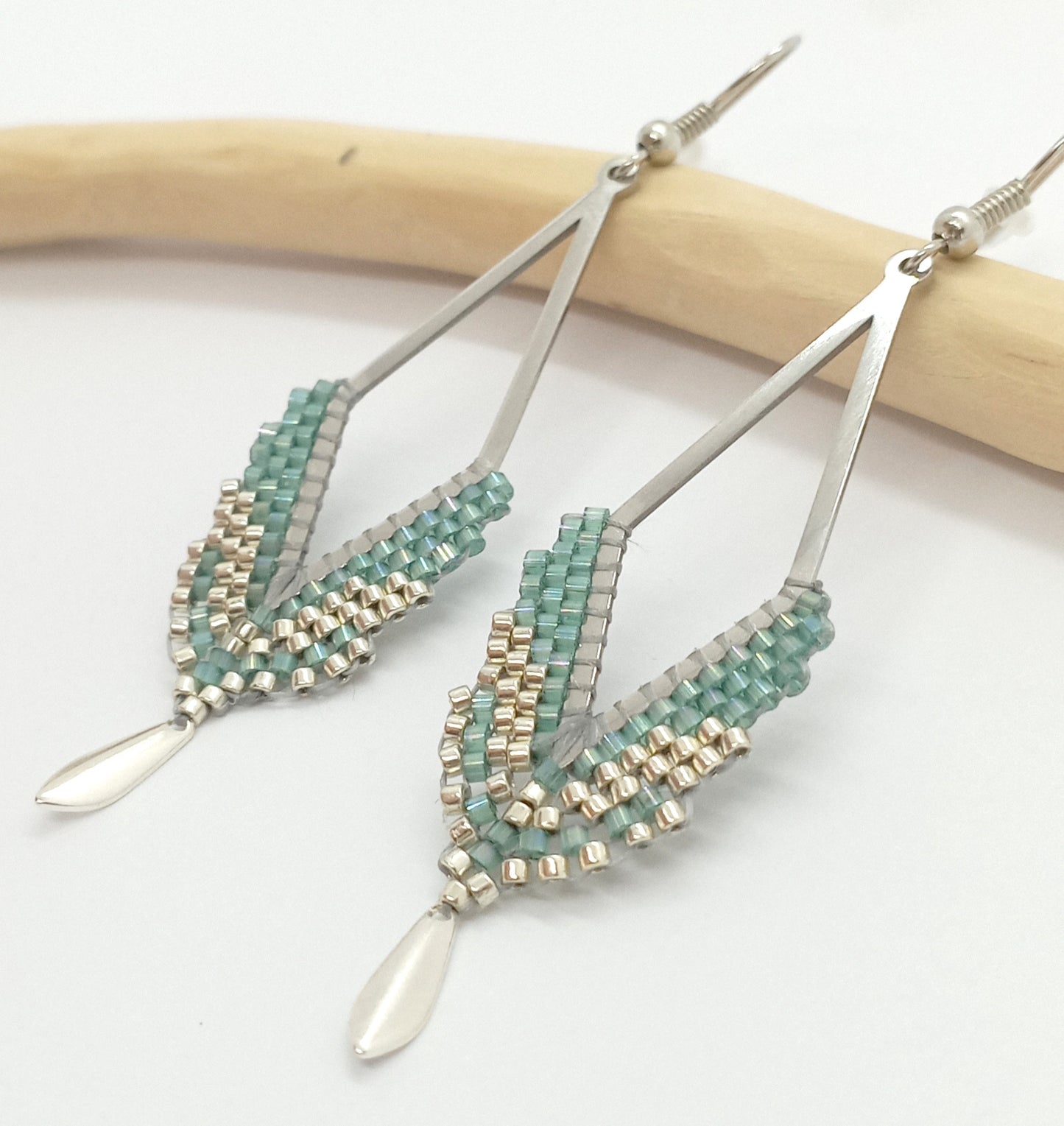 Boucles d'oreilles en forme de losange ajouré, en acier argenté et perles tissées vert d'eau sur un fond blanc et bois clair