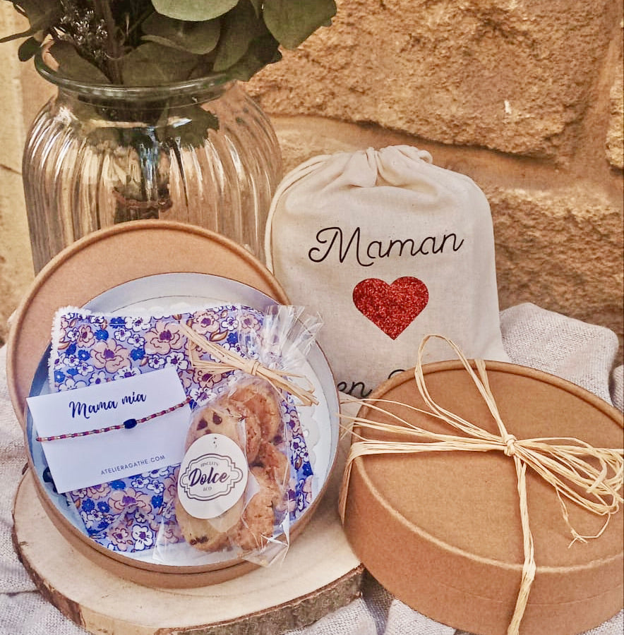 Coffret cadeau avec un bracelet et des gâteaux posé devant un bouquet de fleurs et sur une nappe beige. Un mur en pierre derrière. 