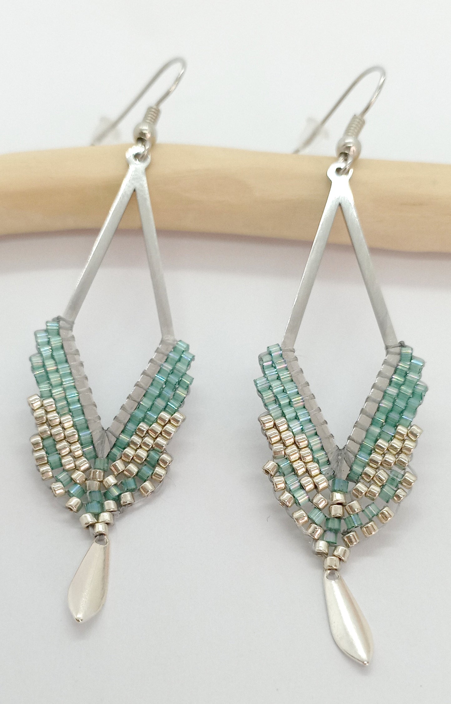 Boucles d'oreilles en forme de losange ajouré en acier argenté et perles miyuki tissées avec un sequin argenté, sur fond blanc et bois clair. 