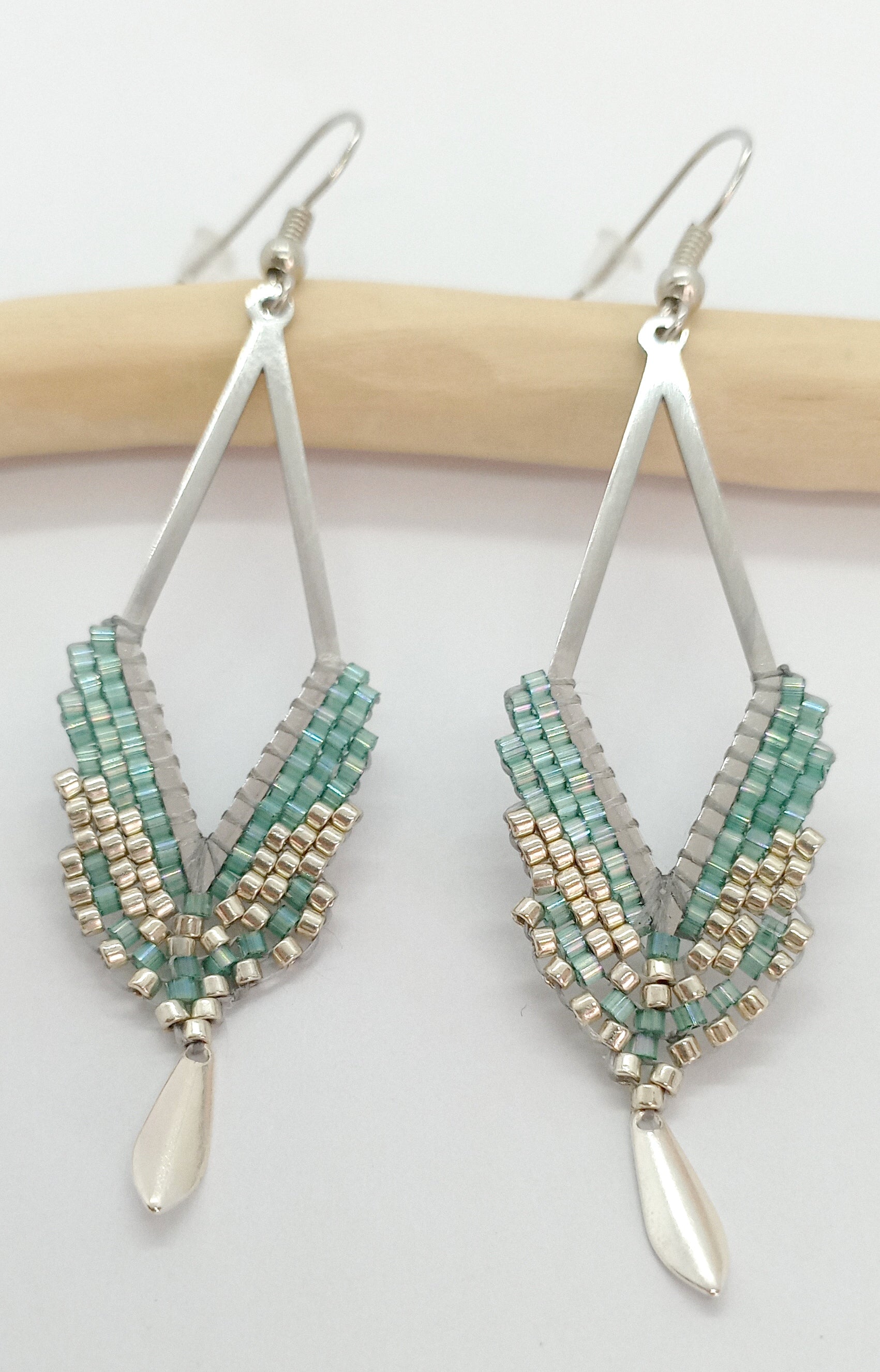 Boucles d'oreilles en forme de losange ajouré en acier argenté et perles miyuki tissées avec un sequin argenté, sur fond blanc et bois clair. 